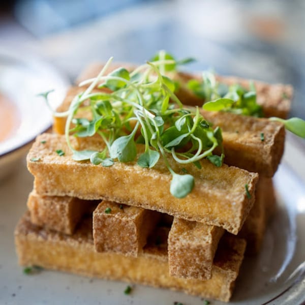 Crispy golden-brown tofu fries, stacked and garnished with fresh green microgreens on a white plate.