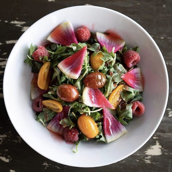 A vibrant salad with arugula, watermelon radishes, raspberries, and cherry tomatoes in a white bowl.