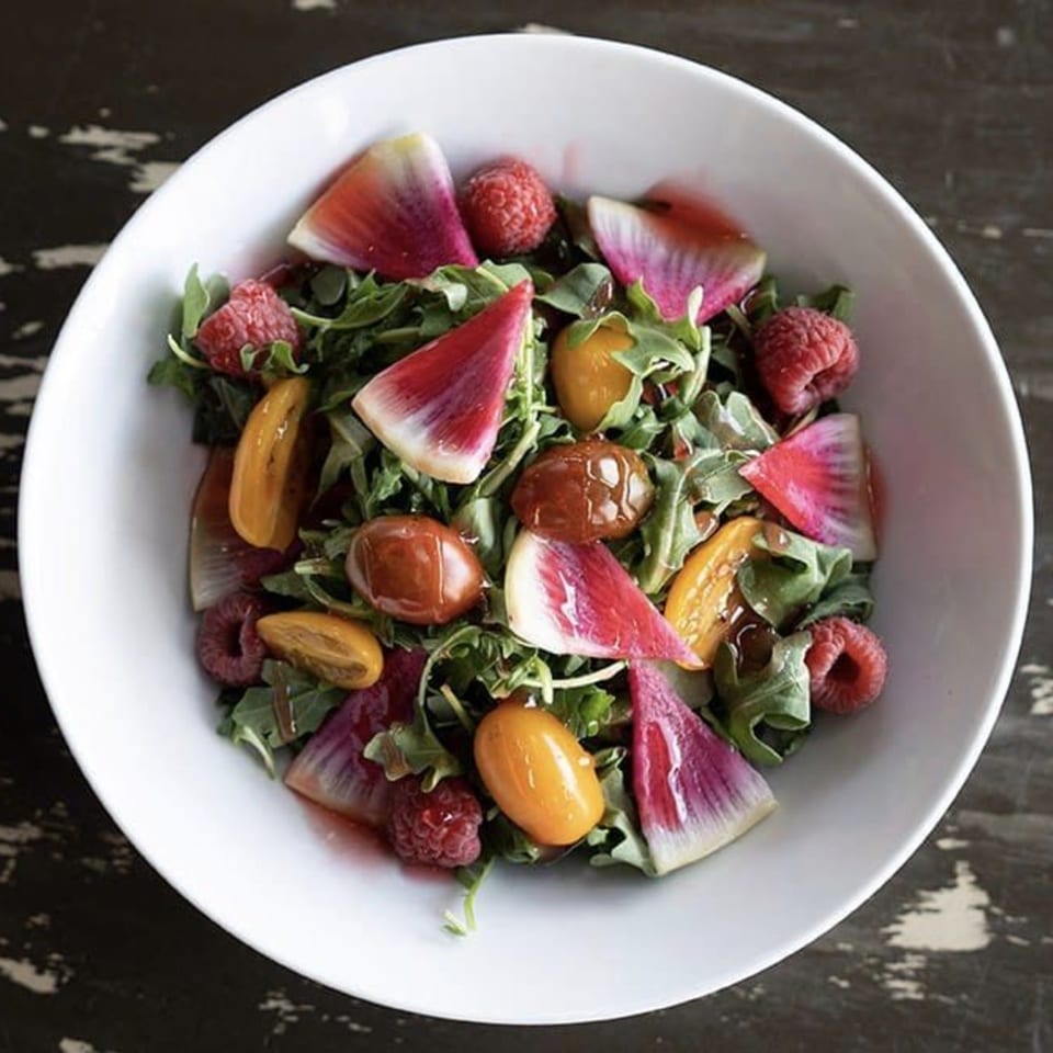 A vibrant salad with arugula, watermelon radishes, raspberries, and cherry tomatoes in a white bowl.
