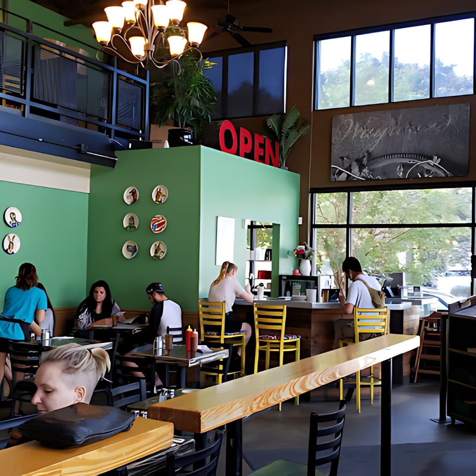 The Wayward Cafe's interior, with customers dining at tables and a counter, featuring green walls and large windows.