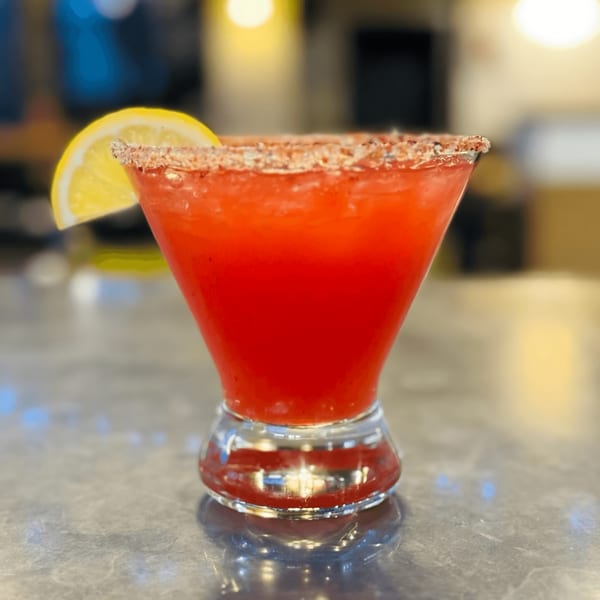 A vibrant red cocktail in a martini glass, garnished with a lemon slice and a sugar-salt rim.