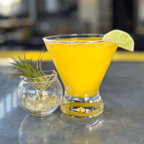 A vibrant yellow cocktail with a lime wedge, next to a small glass with an air plant.