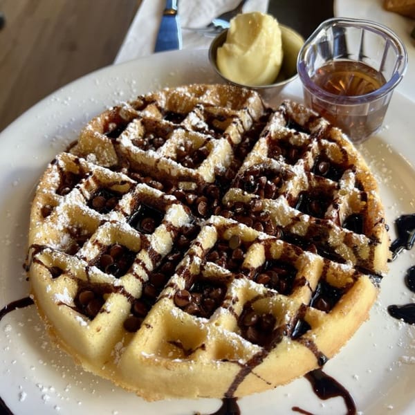 A chocolate chip waffle with powdered sugar, chocolate drizzle, butter, and syrup on a white plate.