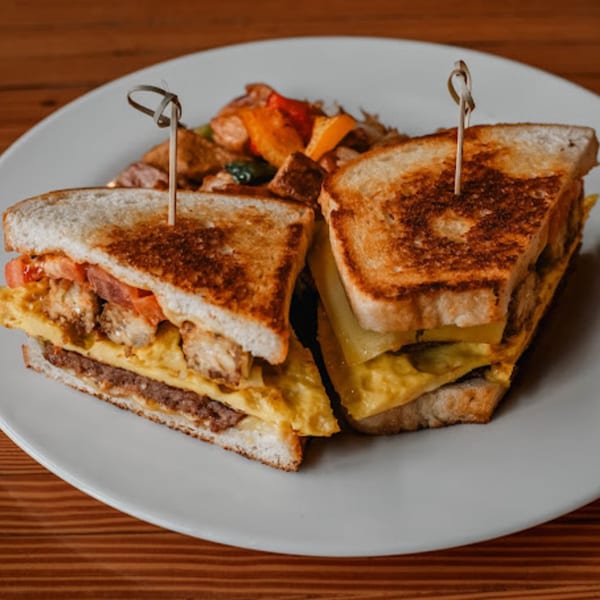 A grilled breakfast sandwich with egg, sausage, and vegetables, served with roasted potatoes on a white plate.